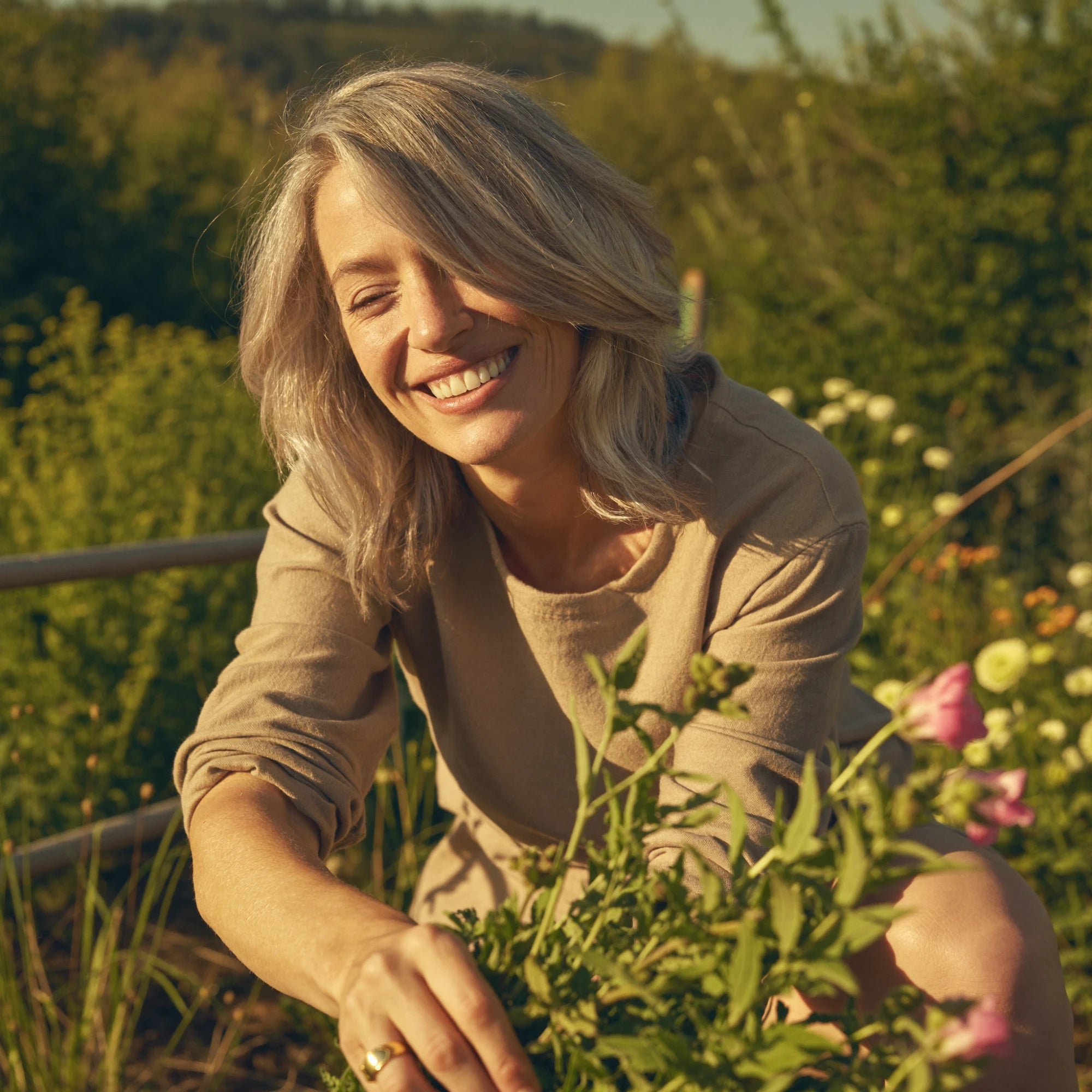 A smiling woman kneels in a sunny garden tending flowers, wearing a gold EverCalm ring.